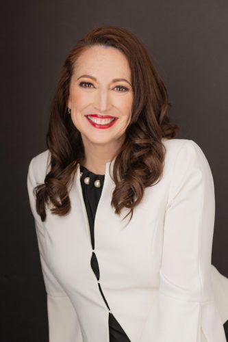 Headshot photo of Eliz Greene wearing a white jacket in front of a dark gray background
